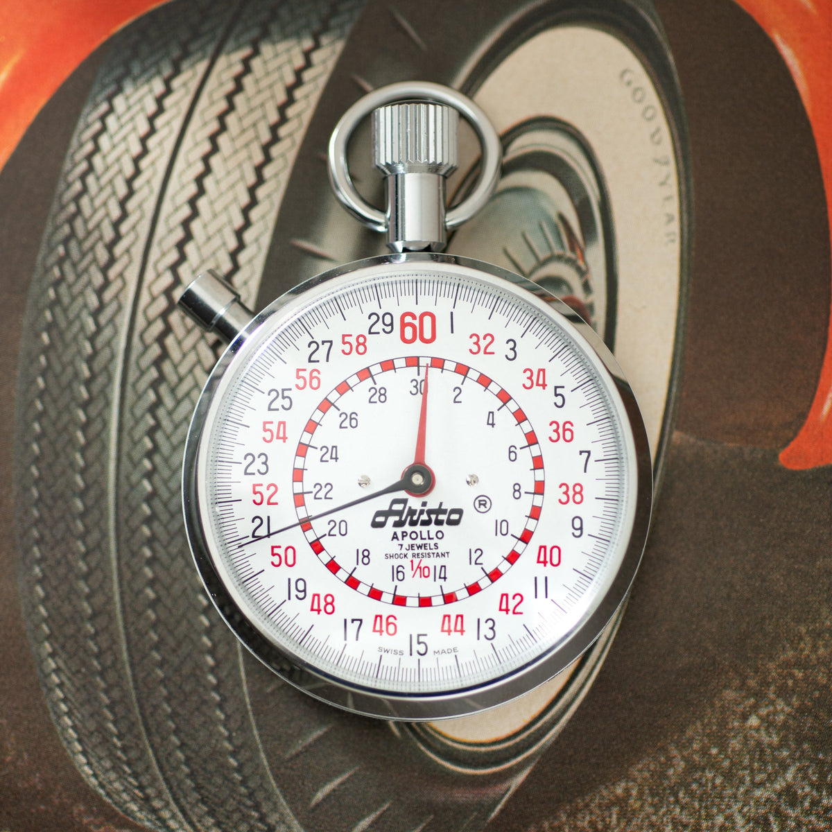 Aristo Apollo stopwatch with white dial, red numerals, and chrome case displayed on vintage tire background.