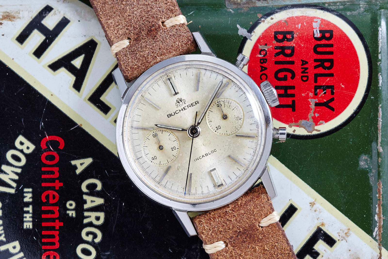 A Bucherer Chronograph with a beige dial and brown leather strap rests atop a vintage “HALF AND HALF” BURLEY AND BRIGHT tin—Swiss luxury meets classic nostalgia.