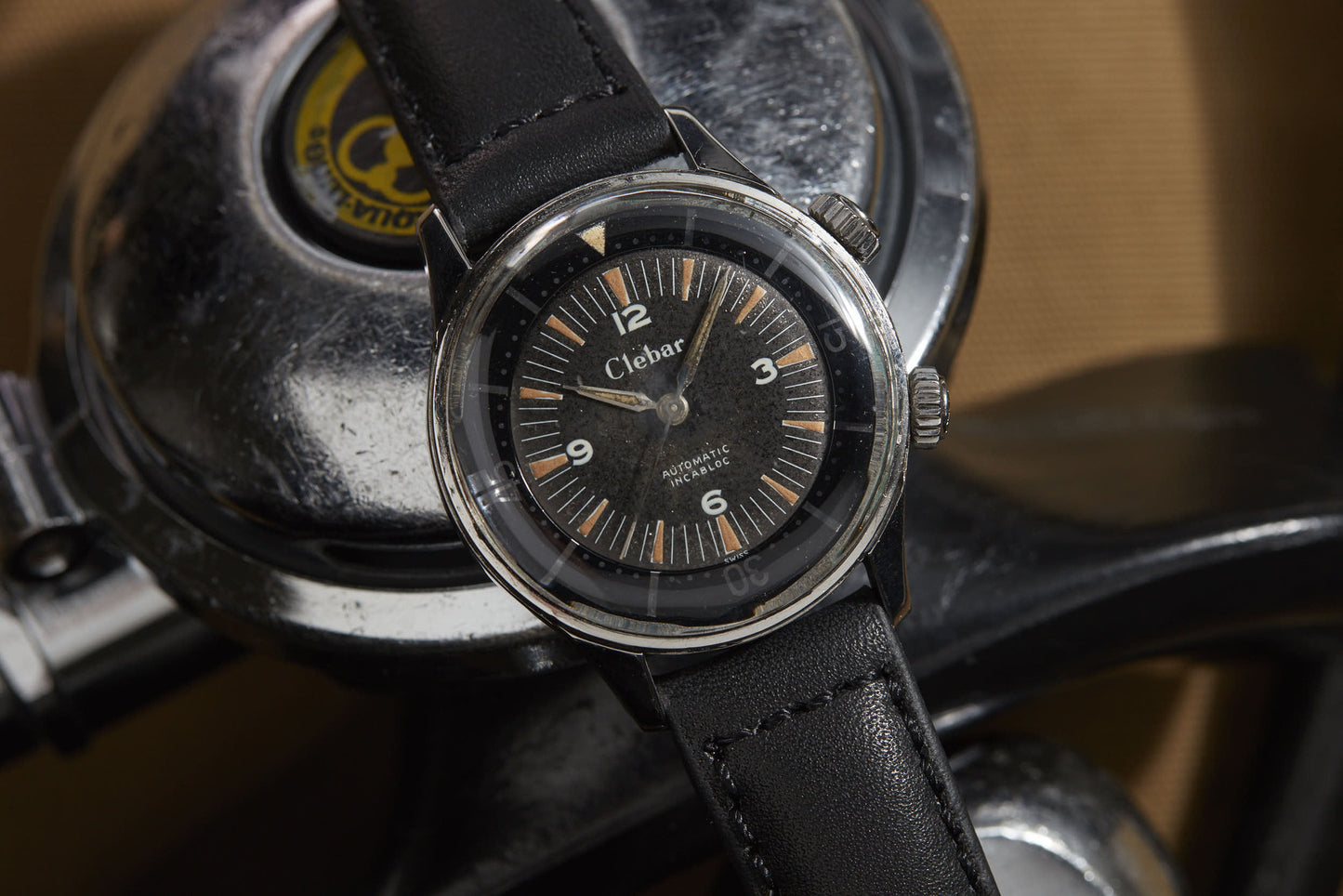 A Clebar Super Compressor wristwatch with a black leather strap and black dial sits on a metallic surface, evoking diving suit precision, with a circular Qualitron label in the background.