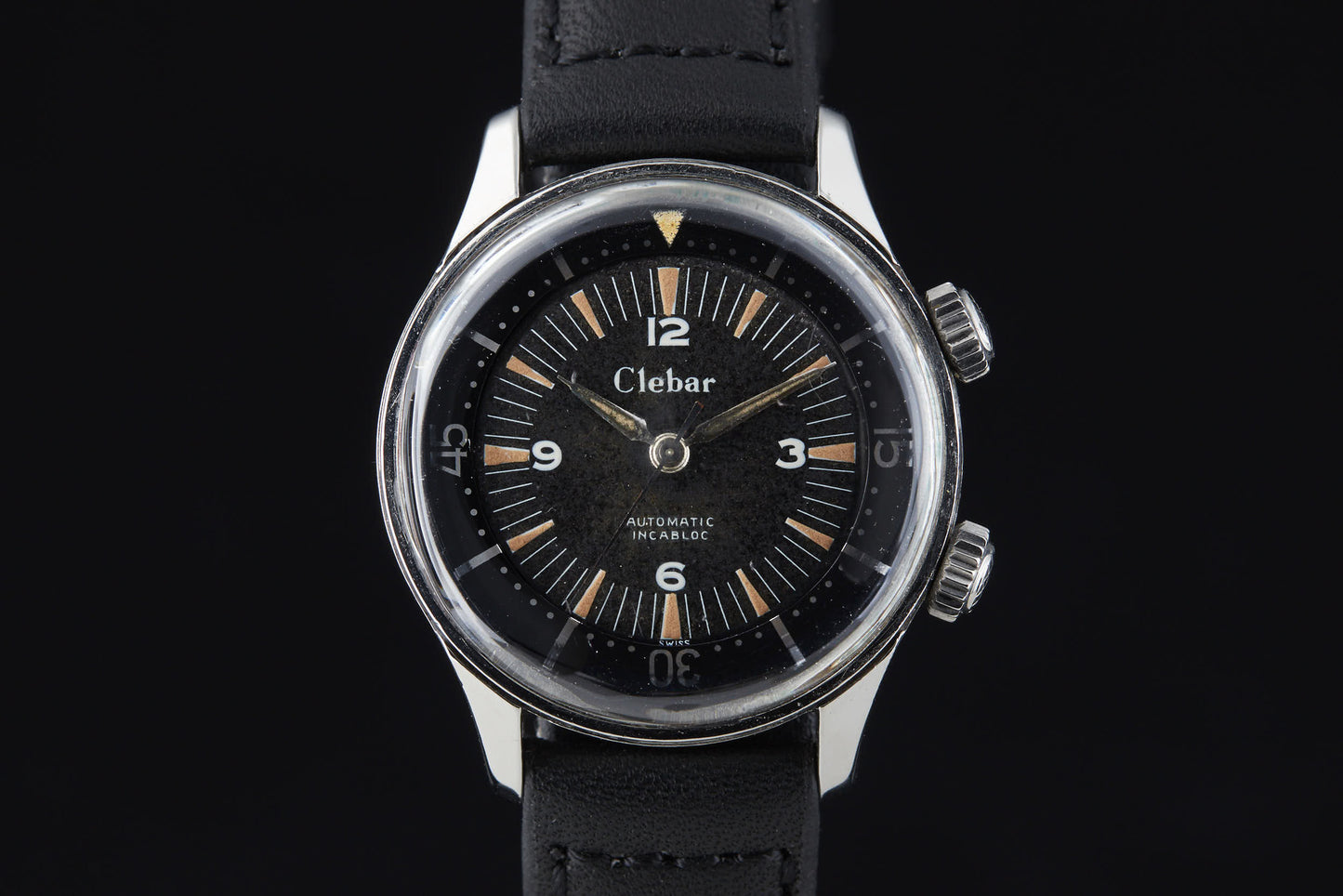 The Clebar Super Compressor by Clebar features a black dial with white numerals, two crowns, a stainless steel case, and a black leather strap—evoking classic diving gear—set against an elegant black background.