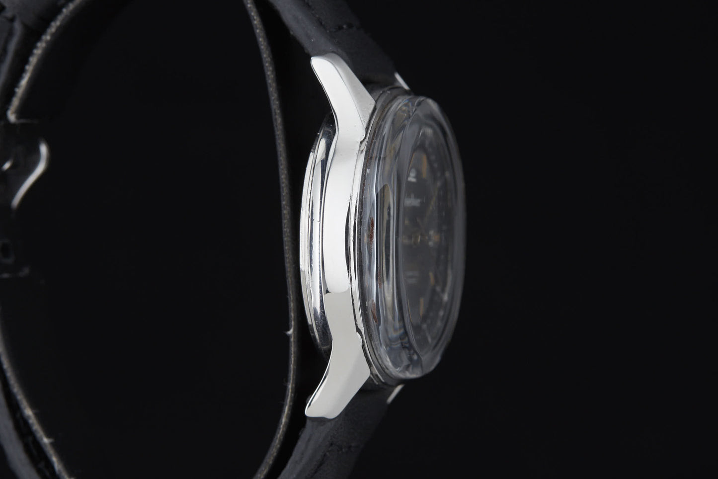 A close-up side view of the Clebar Super Compressor by Clebar showcases its round silver case and black leather strap, set against a black background. The watch face is partially visible through the clear crystal, evoking the sleek precision of a pressure regulator.