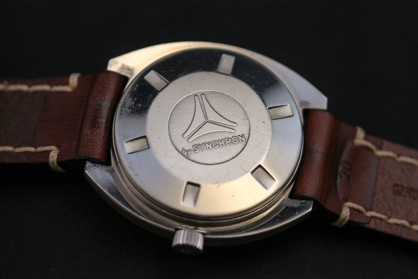 Close-up of the back of a DOXA Sub 300T Sharkhunter Aqua Lung wristwatch with a brown leather strap. The metal case features an engraved logo of three intersecting lines above "by SYNCHRON," evoking an iconic divers' design.
