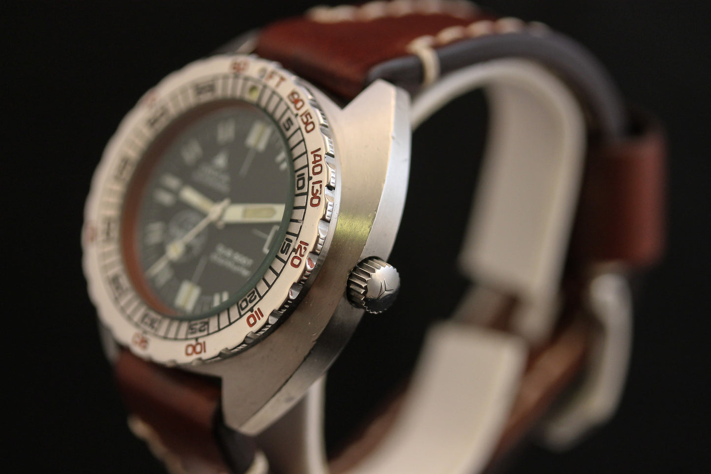 A close-up of the DOXA Sub 300T Sharkhunter Aqua Lung dive watch with a brown leather strap, silver bezel with red markings, and dark dial. The iconic diver’s watch is angled to display the crown against a black background.