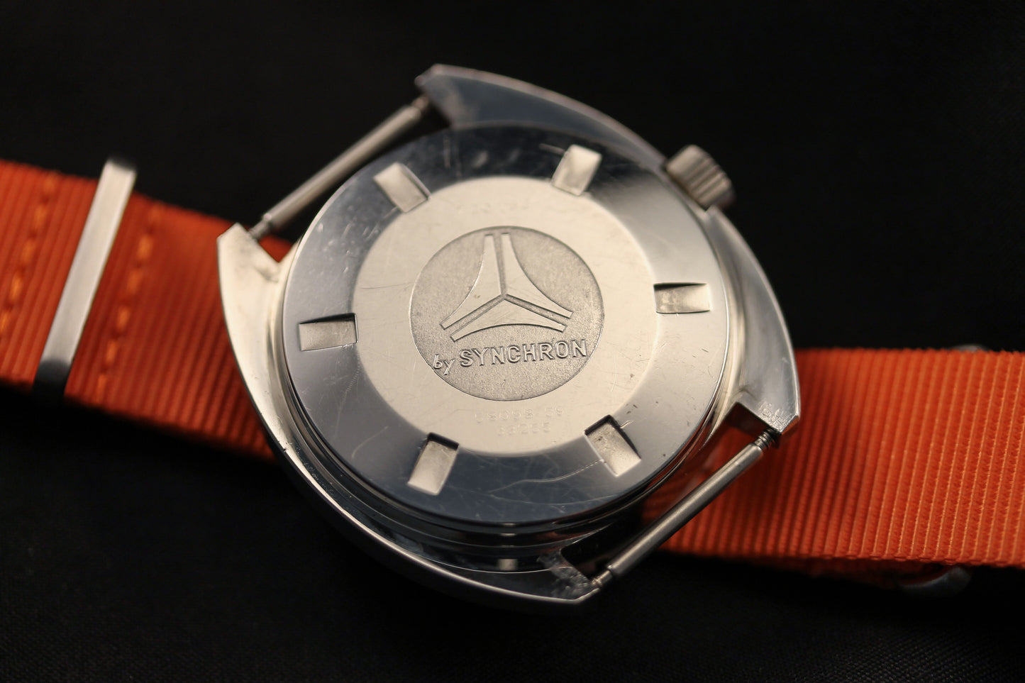 A close-up of the DOXA Sub 300T Sharkhunter diver's watch with an orange strap, featuring the engraved Synchron logo and Sub Series markings on the case back against a black background.