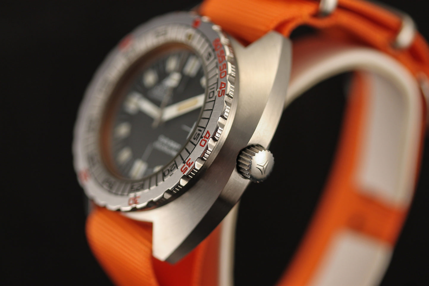 Close-up of the DOXA Sub 300T Sharkhunter diver’s watch, featuring a black dial, silver bezel with red numbers, and an orange strap. The sharply focused crown and case highlight classic DOXA style against a blurred black background.