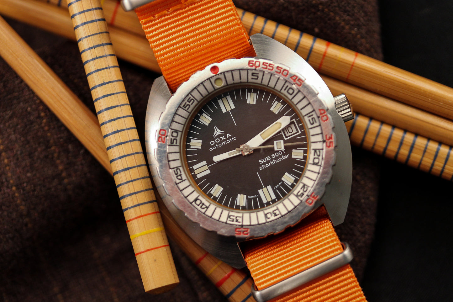 A DOXA Sub 300T Sharkhunter diver's watch with a stainless steel case and orange strap rests on brown fabric, surrounded by bamboo rods with red, yellow, and blue markings. The bold black dial features striking white indices.