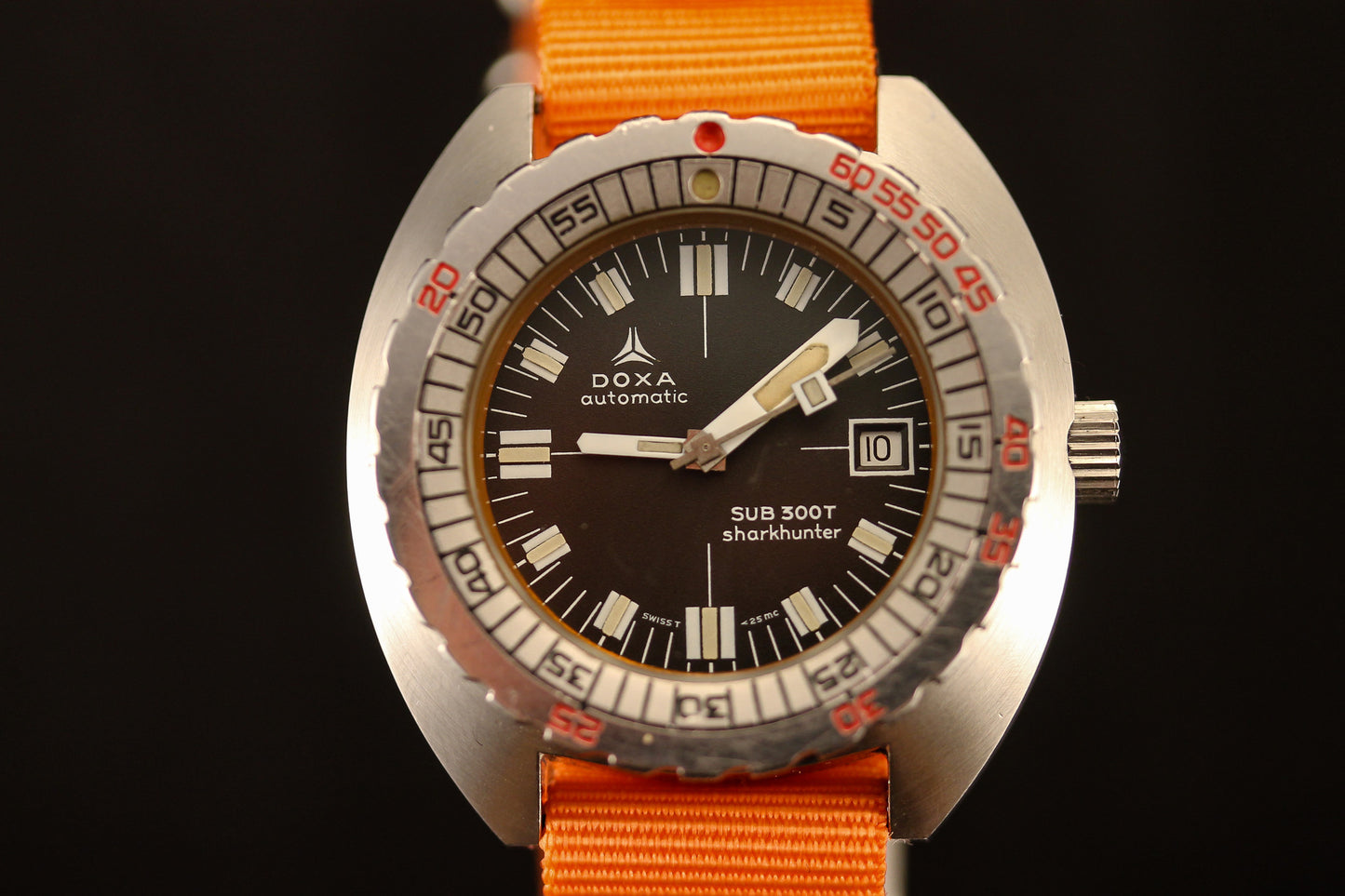 A close-up of the DOXA Sub 300T Sharkhunter by DOXA, featuring a black dial with bold white indices, silver bezel with red and black markings, orange NATO strap, and a date window at 3 o’clock.