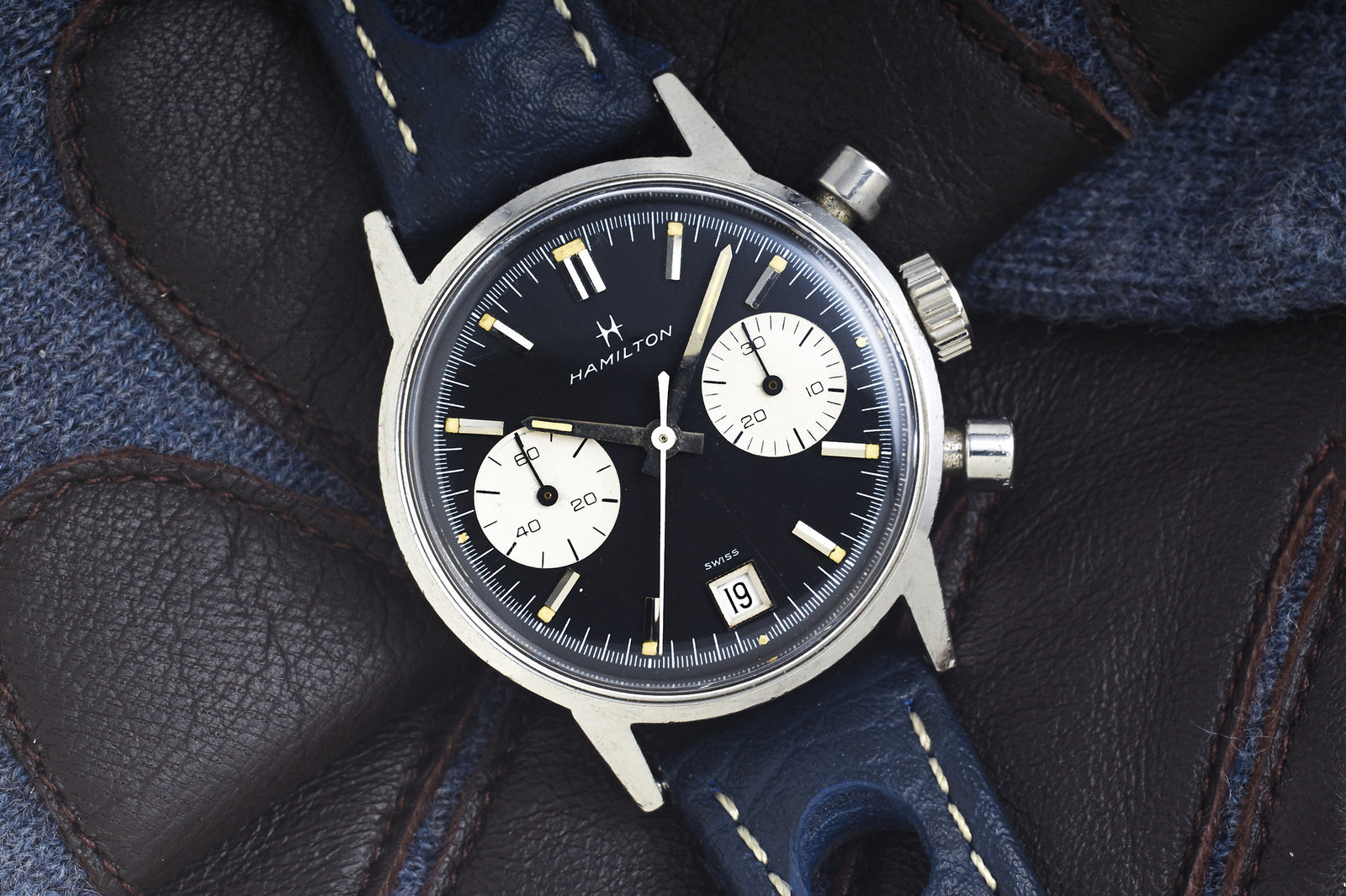 A close-up of the Hamilton "Poor Man's Carrera" Dato reveals its black dial, two white subdials, a date window at 6 o’clock, and a blue leather strap resting on dark leather gloves.