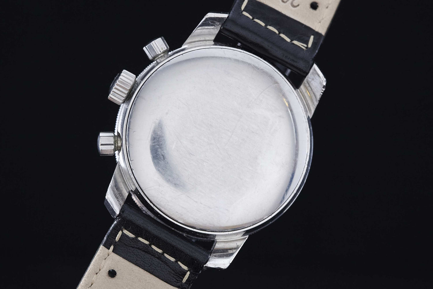 A close-up of the Heuer Autavia, a manually-wound chronograph by Heuer, featuring a plain silver case, black leather strap with white stitching, and three metal pushers, inspired by vintage motor sports watches.