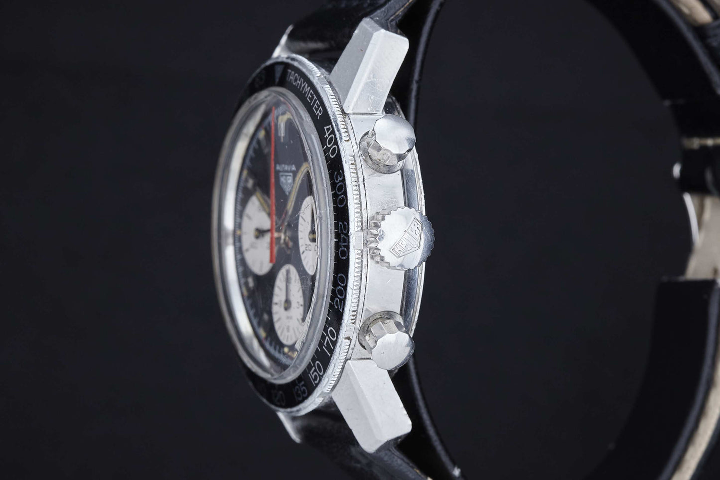 A close-up side view of the Heuer Autavia by Heuer featuring a black bezel, silver pushers, and black leather strap. Its multi-subdial face highlights the model’s legacy in motorsports, set against a dark background.