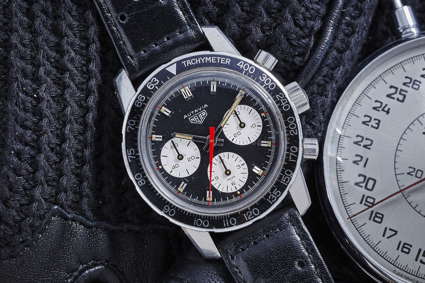 A close-up of the Heuer Autavia by Heuer, a motorsports classic with a black dial, three white subdials, red seconds hand, black leather strap, and tachymeter bezel on a knitted black surface beside a stopwatch.