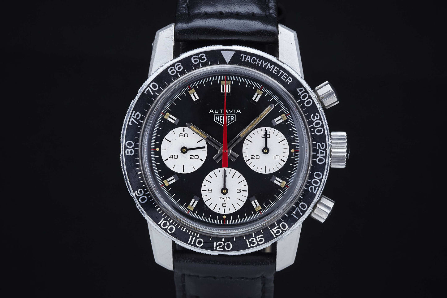 The Heuer Autavia by Heuer is a vintage chronograph with a black dial, three white subdials, tachymeter bezel, red second hand, and black leather strap, displayed against a black background.