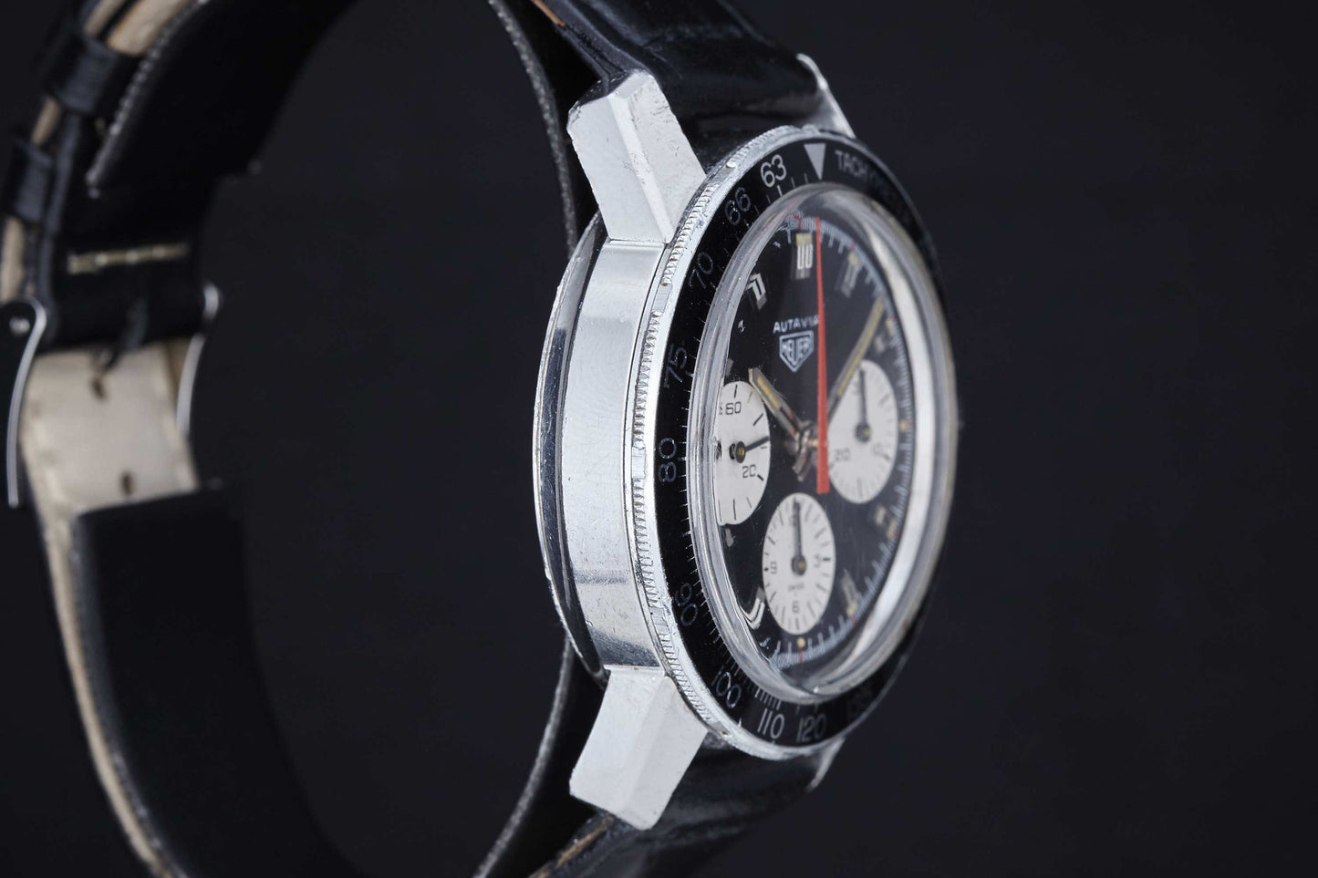 A close-up side view of the Heuer Autavia by Heuer, a manual chronograph admired in motor sports, with a black leather strap, black bezel, and three white subdials on its face set against a dark background.