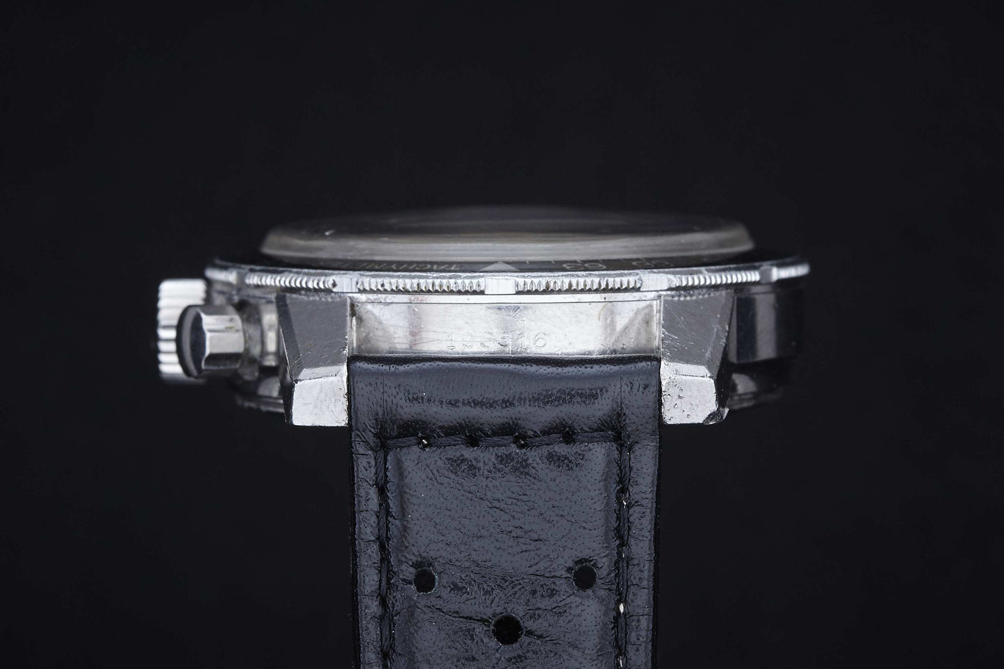 Side view of the Heuer Autavia by Heuer, featuring a black leather strap, visible crown, and textured bezel—an iconic motor sports watch—displayed against a plain black background.