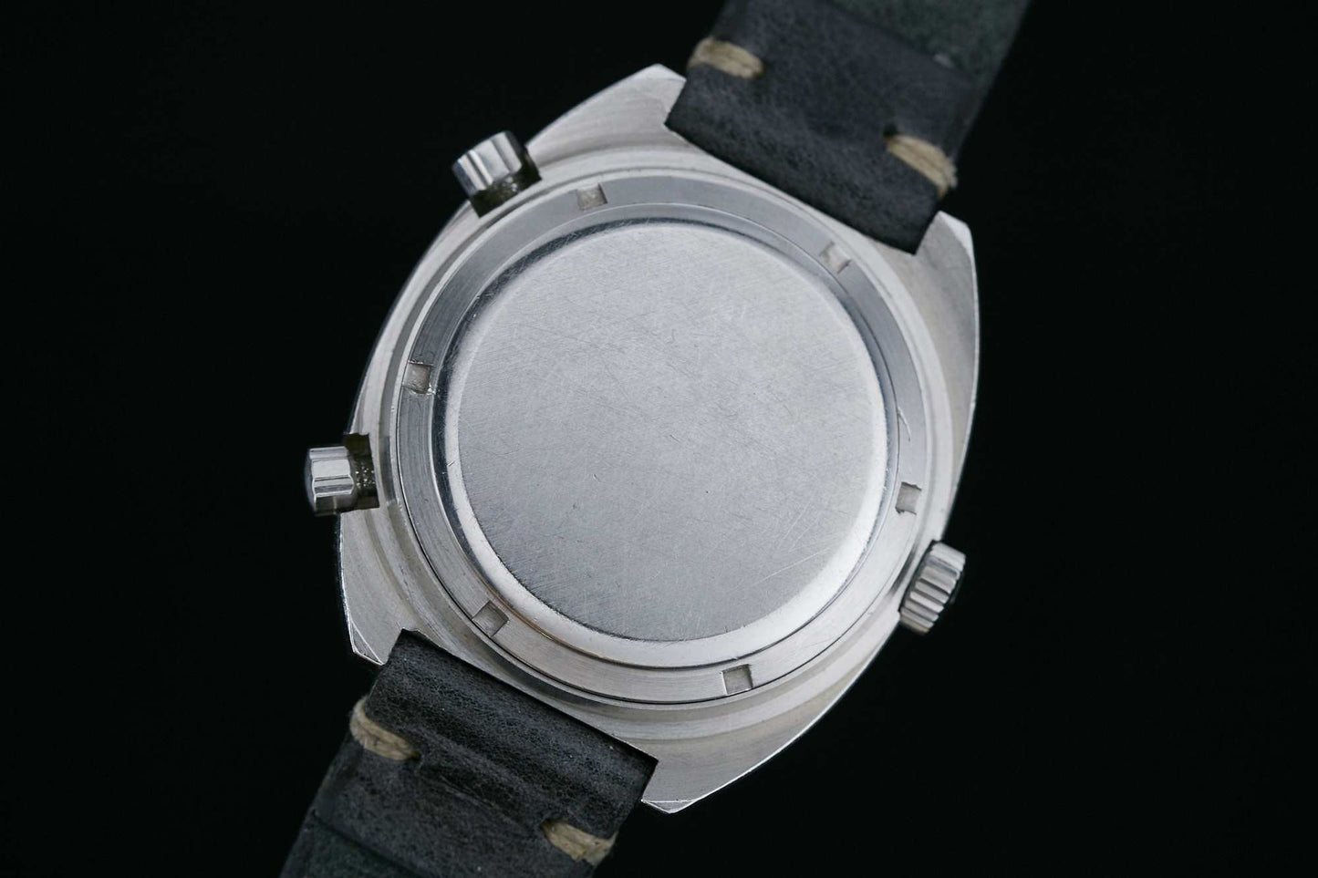A close-up of the back of a silver Heuer Autavia GMT automatic chronograph case with three crowns and a black leather strap, set against a black background. The plain case back has no visible engravings or markings.