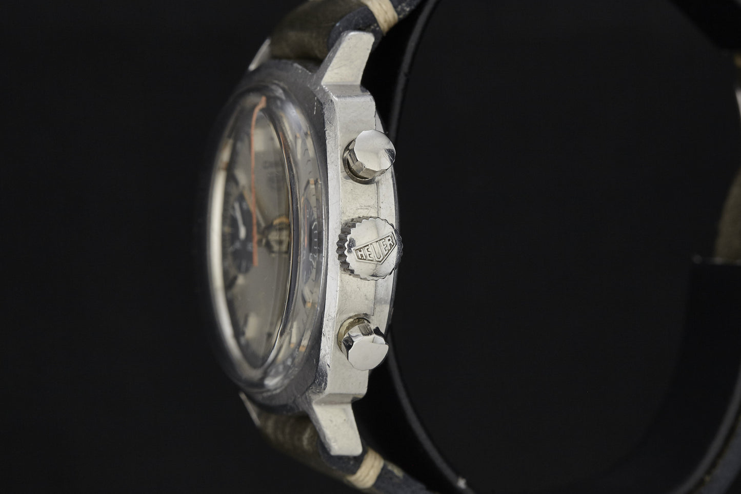Close-up side view of the Heuer Camaro wristwatch, highlighting the case, two pushers, and a crown engraved with the iconic Heuer logo—all set against a black background.