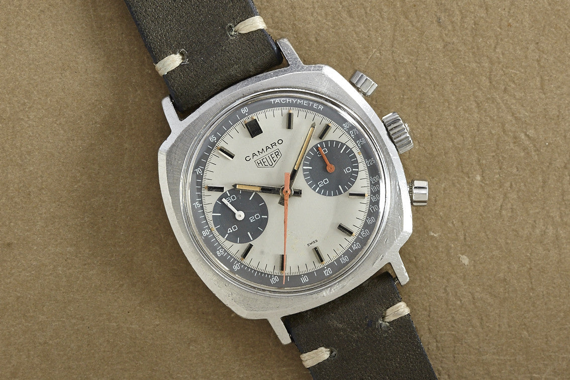 Discover the Heuer Camaro by Heuer—a vintage wristwatch with a silver dial, two black subdials, tachymeter scale, and a black leather strap with white stitching. Elegantly presented on a brown textured background.