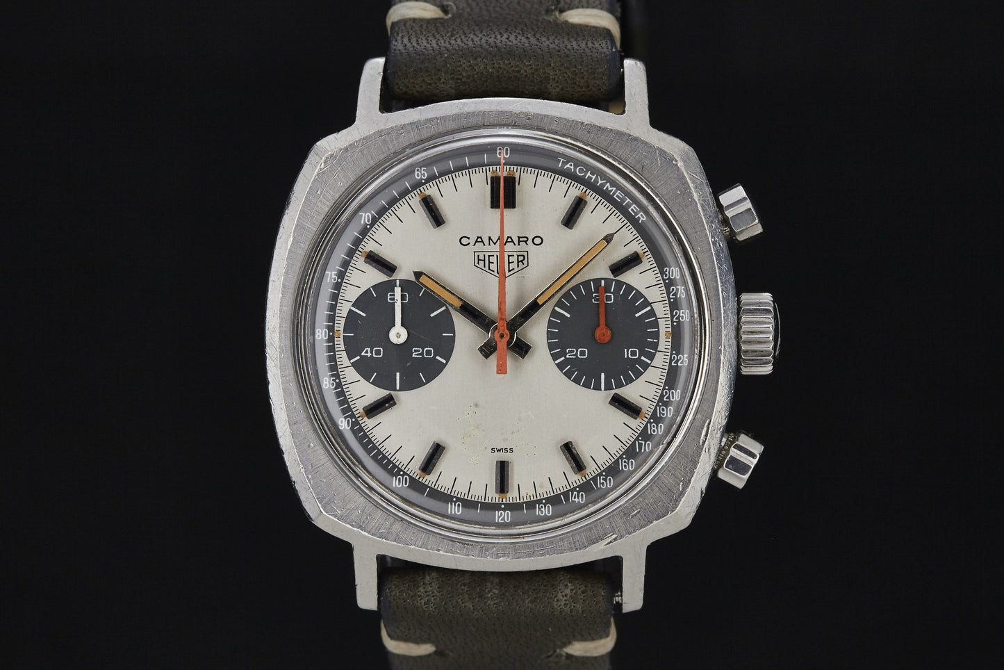 The Heuer Camaro by Heuer features a silver square case, two black subdials, tachymeter scale, and a sleek black leather strap, all set against a striking black background.