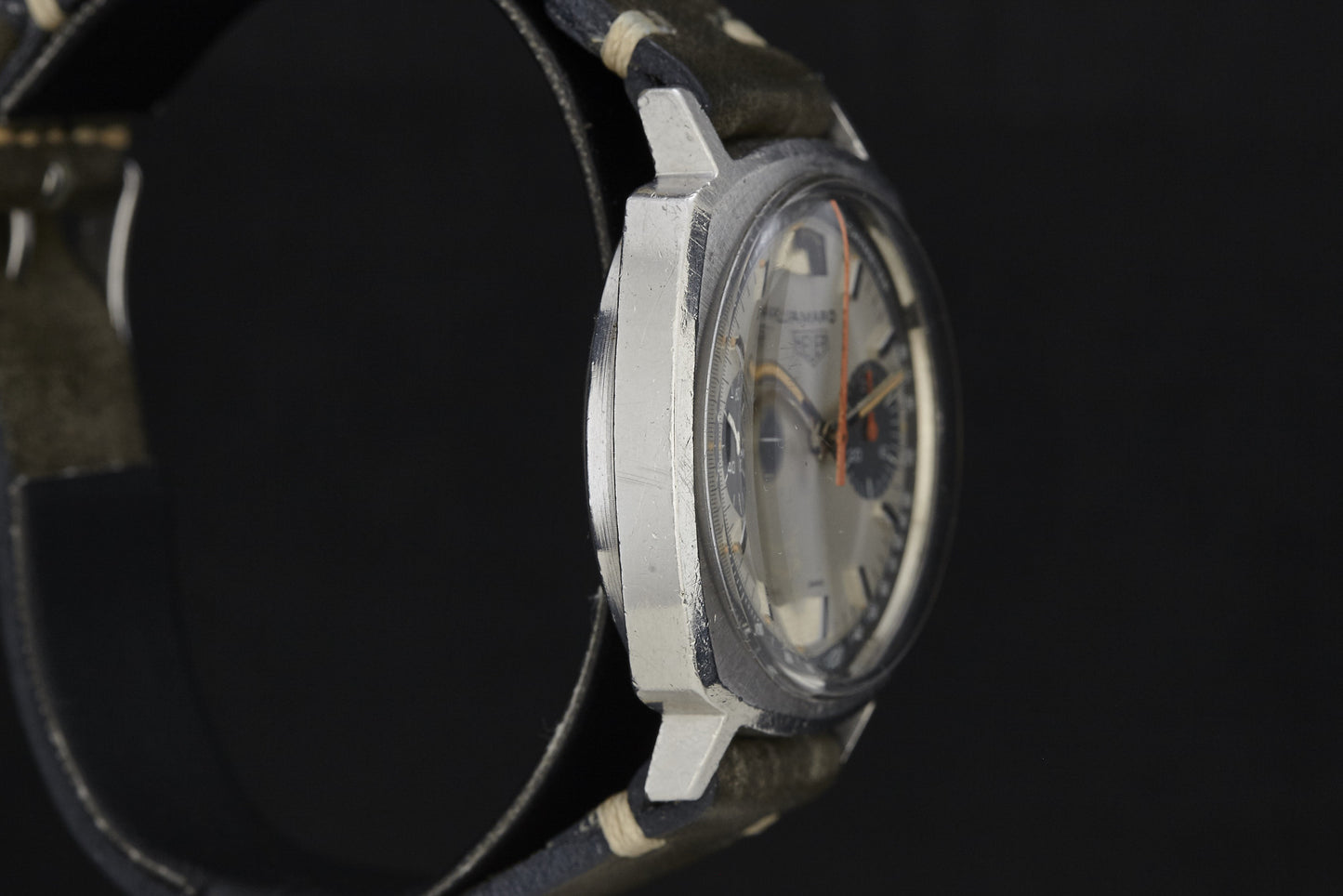 A close-up side view of a vintage Heuer Camaro wristwatch with a scratched silver metal case, black leather strap, and a partially visible dial set against a dark background.
