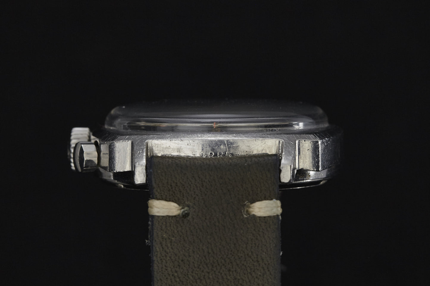 A close-up side view of the Heuer Camaro by Heuer showcases its vintage metal case, prominent crown, and aged greenish-brown leather strap against a black background.