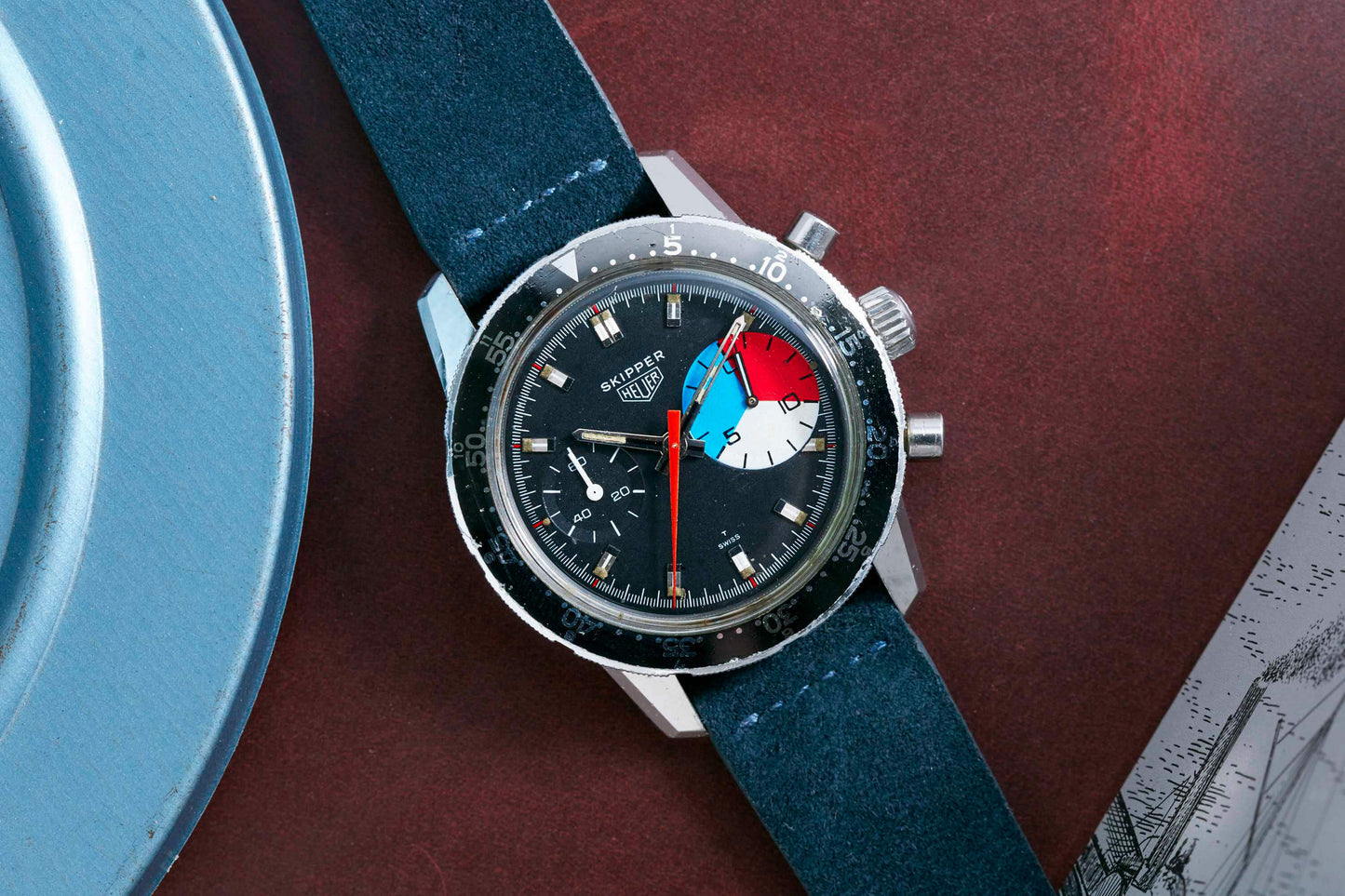 The Heuer Skipper Reference 7764 wristwatch by Heuer, featuring a dark blue strap and a colorful blue, red, and white subdial, rests on a reddish surface beside a blue dish. Detailed Description Coming Soon!.