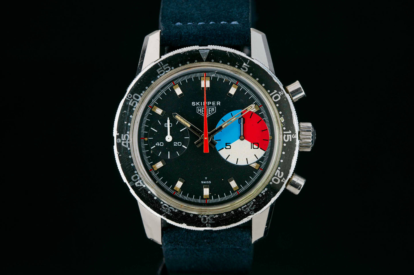 A close-up of a vintage Heuer Skipper Reference 7764 wristwatch with a black bezel, blue strap, and multicolored sub-dial (blue, white, red) set against a black background. Detailed description coming soon!.