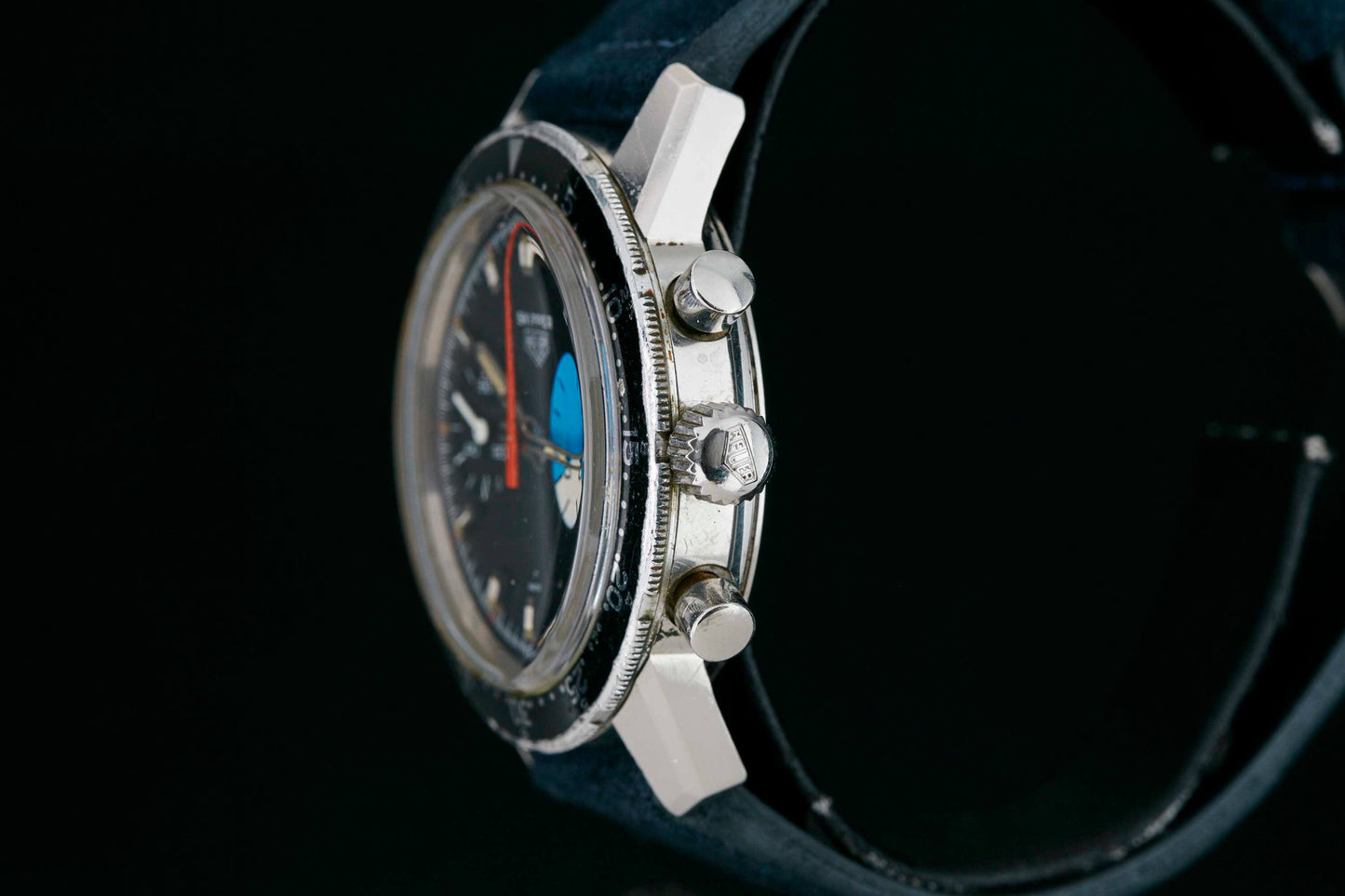 A close-up side view of the Heuer Skipper Reference 7764 wristwatch by Heuer, featuring a silver metal case with two pushers and a crown, black strap, and dark dial with blue accents against a black background. Detailed description coming soon!.