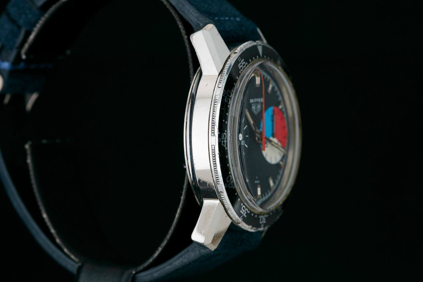 A close-up side view of the Heuer Skipper Reference 7764 by Heuer, featuring a round silver case, black bezel, dark strap, and a black dial with blue and red segments accented by white hour markers. Detailed Description Coming Soon!.