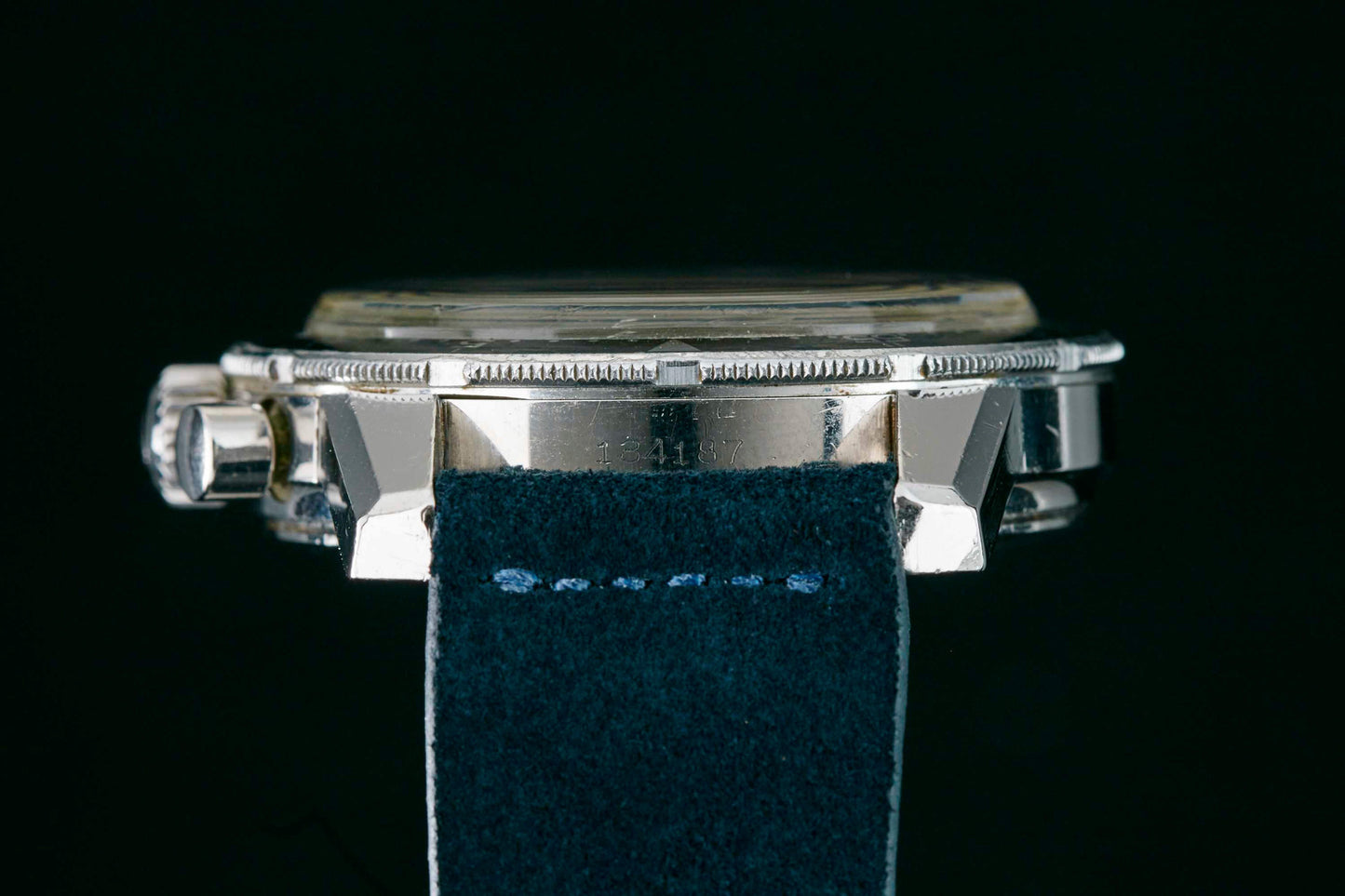 Close-up side view of the Heuer Skipper Reference 7764 by Heuer, featuring a silver case with a dark blue suede strap. Fine bezel and crown details are highlighted against a black background. Detailed description coming soon!.