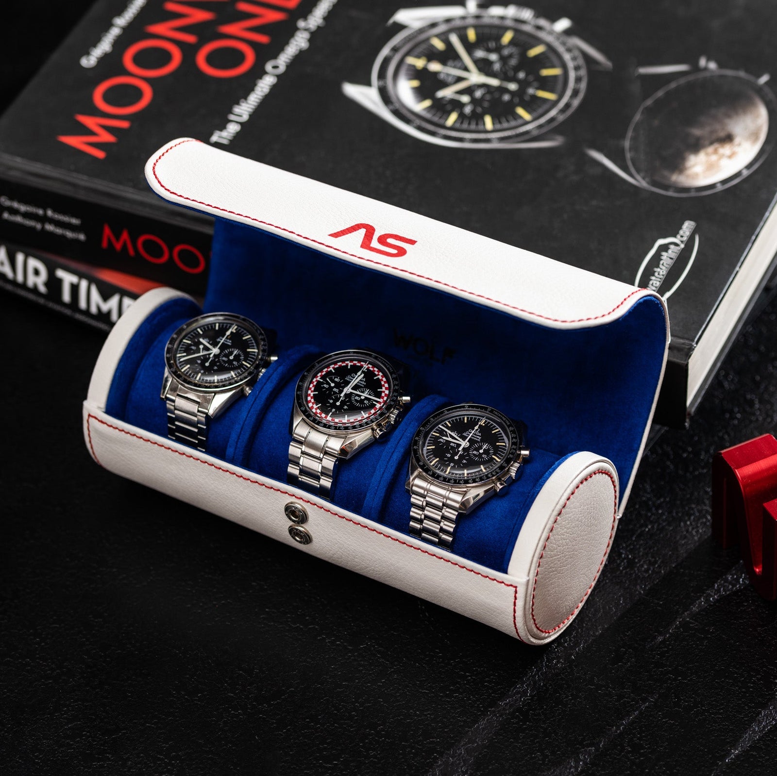 The WOLF Moonshot 3-Piece Travel Tube in white and blue leather holds three silver watches, shown on a dark surface beside Omega Speedmaster books and red decorative letters.