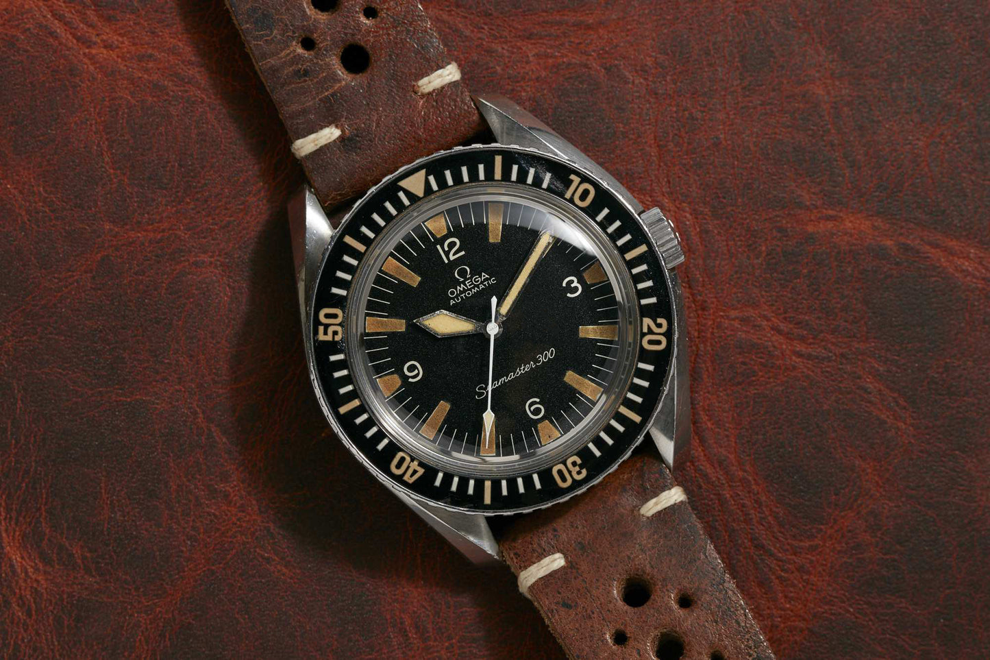 The Omega Seamaster 300 by Omega, featuring a black dial, cream hour markers, and a brown leather strap, rests on textured leather. More details coming soon!.