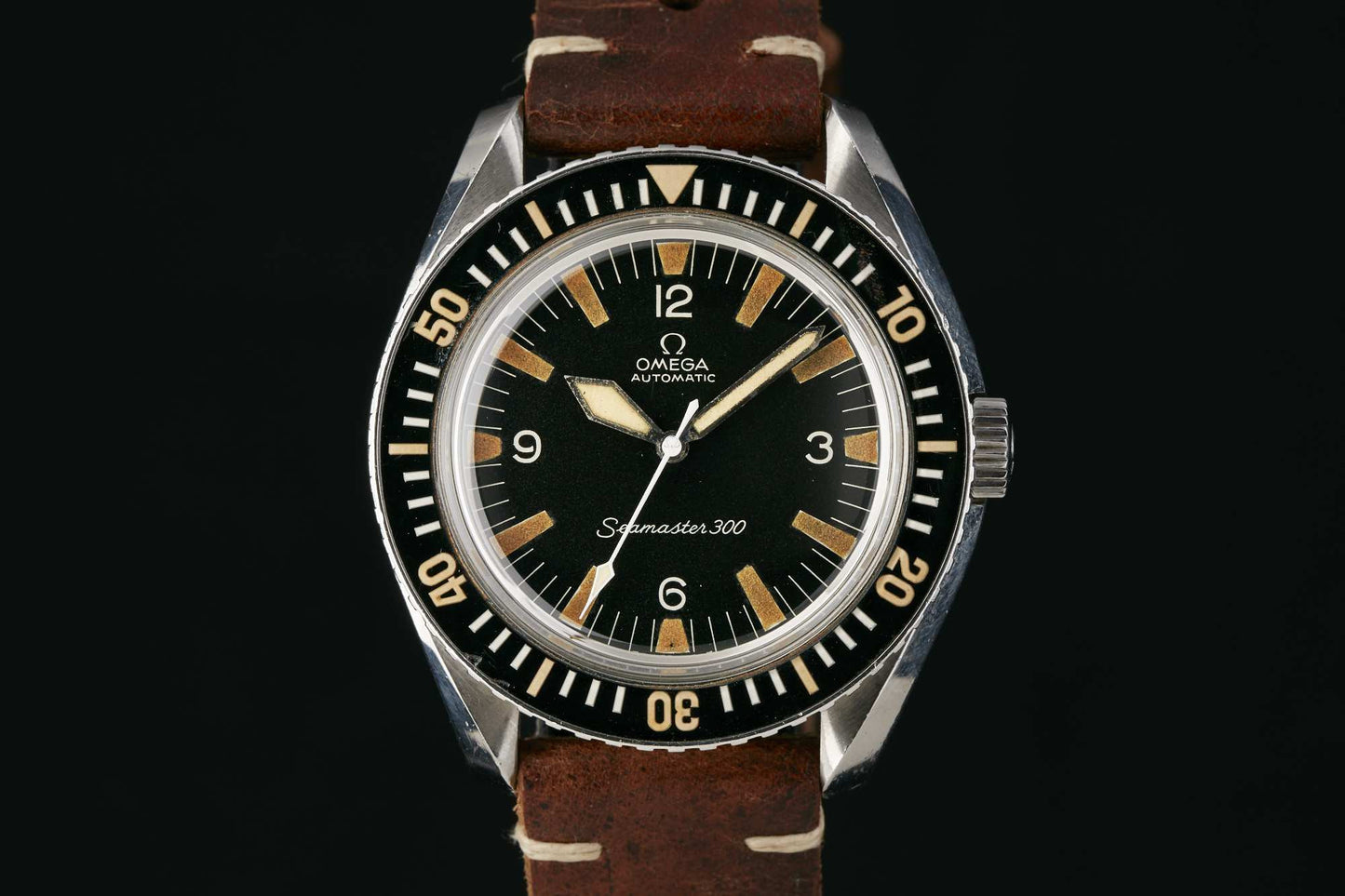 The Omega Seamaster 300 by Omega features a black dial with bold white numerals and indexes, set in a stainless steel case with a rotating bezel and brown leather strap. Detailed description coming soon! Shown on a black background.