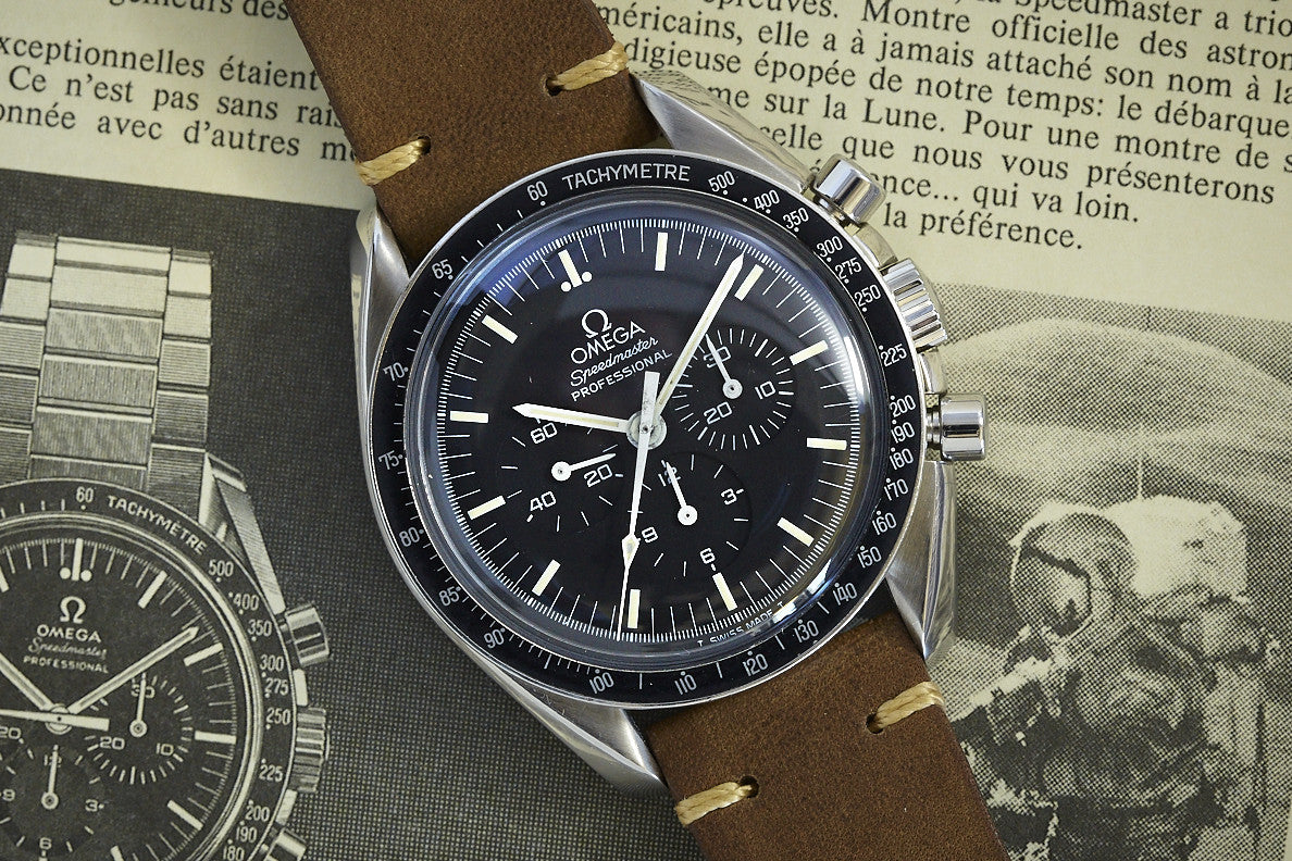 A close-up of the Omega Speedmaster by Omega with a black dial and brown leather strap, set atop a vintage magazine displaying text and an image of a similar NASA chronograph.