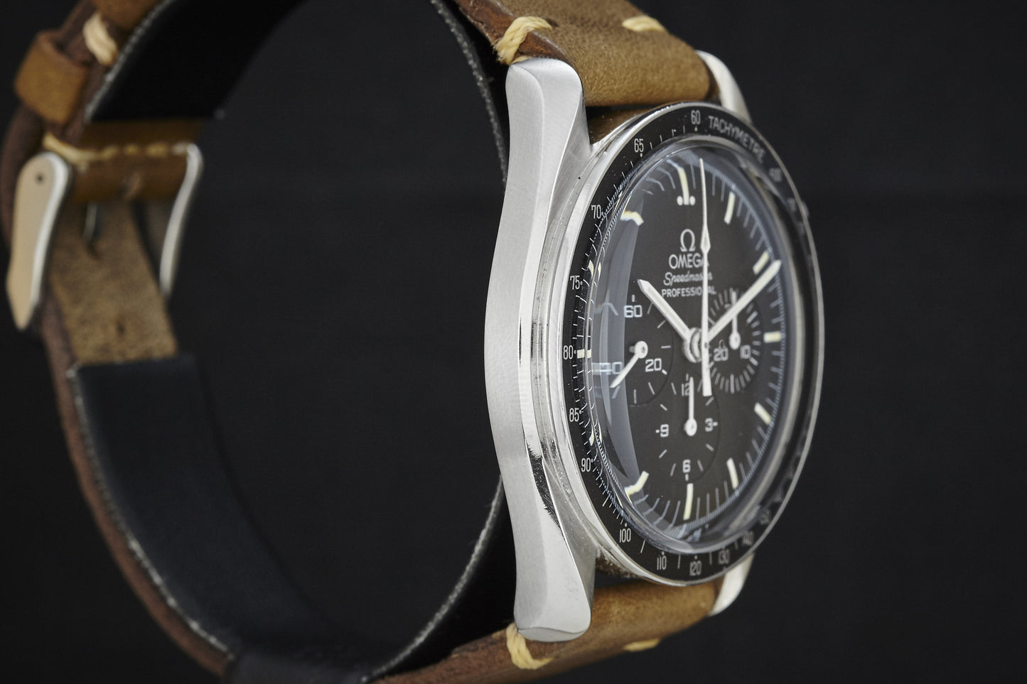 A close-up side view of the Omega Speedmaster by Omega, featuring a black dial, tachymeter bezel, and brown leather strap against a dark background.