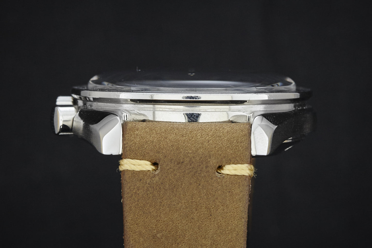 Side view of the Omega Speedmaster by Omega, featuring a polished metal case, flat glass crystal, and tan leather strap with beige stitching set against a black background.