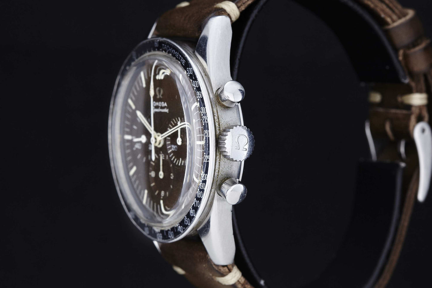 A close-up side view of the Omega Speedmaster Ed White by Omega, featuring a black dial, silver casing, and brown leather strap against a black background. The Omega logo appears on the crown.