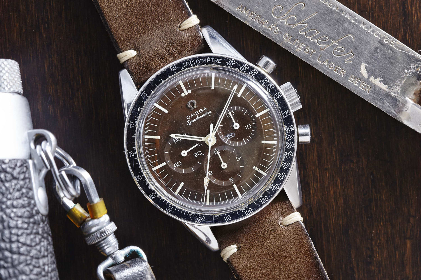 An Omega Speedmaster Ed White by Omega, featuring a brown leather strap, rests on a dark wooden surface beside a Schaefer-engraved metal tool and an item with leather texture and metal clasp.