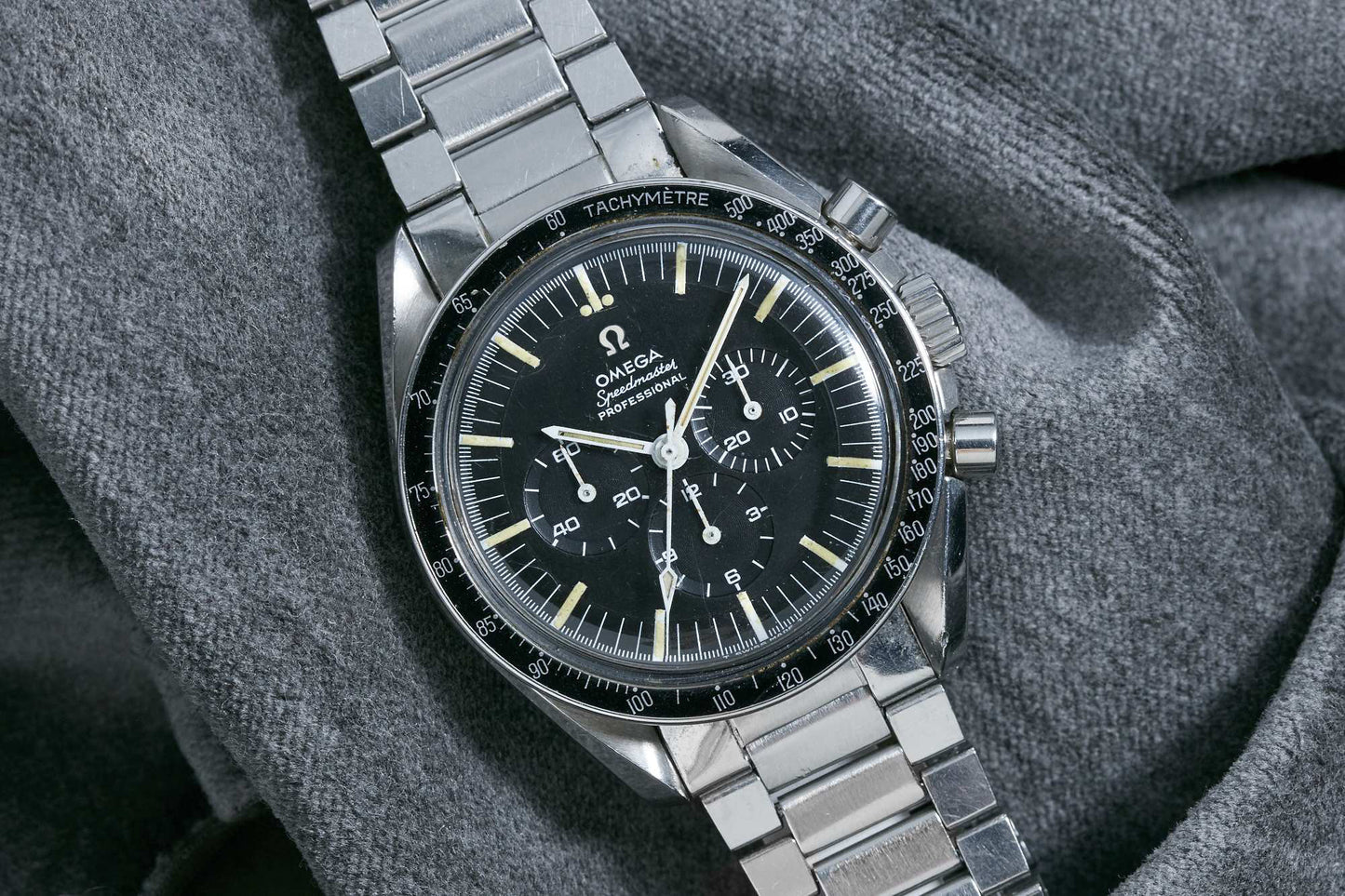 A close-up of an Omega Speedmaster Professional by Omega, featuring a stainless steel bracelet, black dial with three subdials, and a tachymeter bezel, resting on soft gray fabric.