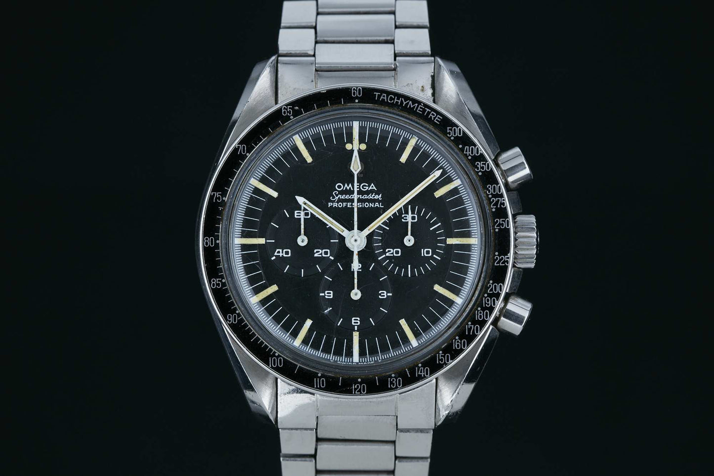 A close-up of the Omega Speedmaster Professional by Omega, featuring a black dial, silver hour markers, three subdials, and a stainless steel bracelet against a black background.