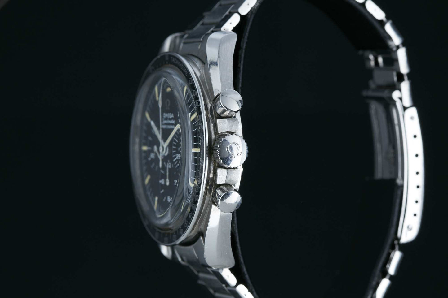A close-up side view of the Omega Speedmaster Professional by Omega displays its detailed crown, two pushers, and partially visible racing chronograph face. The metal bracelet stands out sharply against a black background.