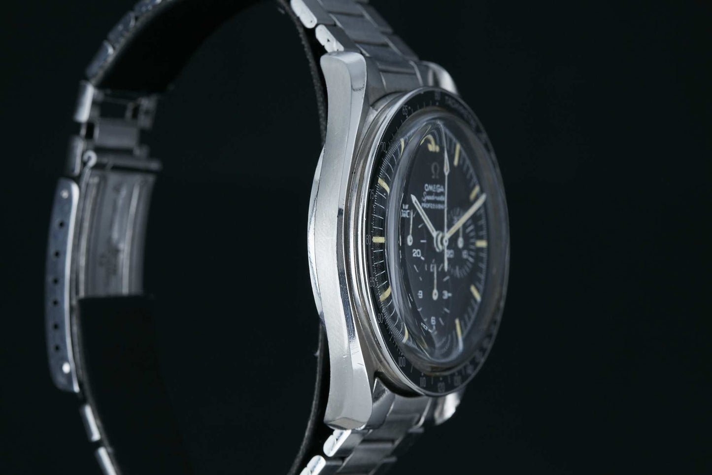 A close-up side view of the Omega Speedmaster Professional by Omega, featuring a stainless steel case, black dial, silver hour markers, and a metal link bracelet against a dark background.