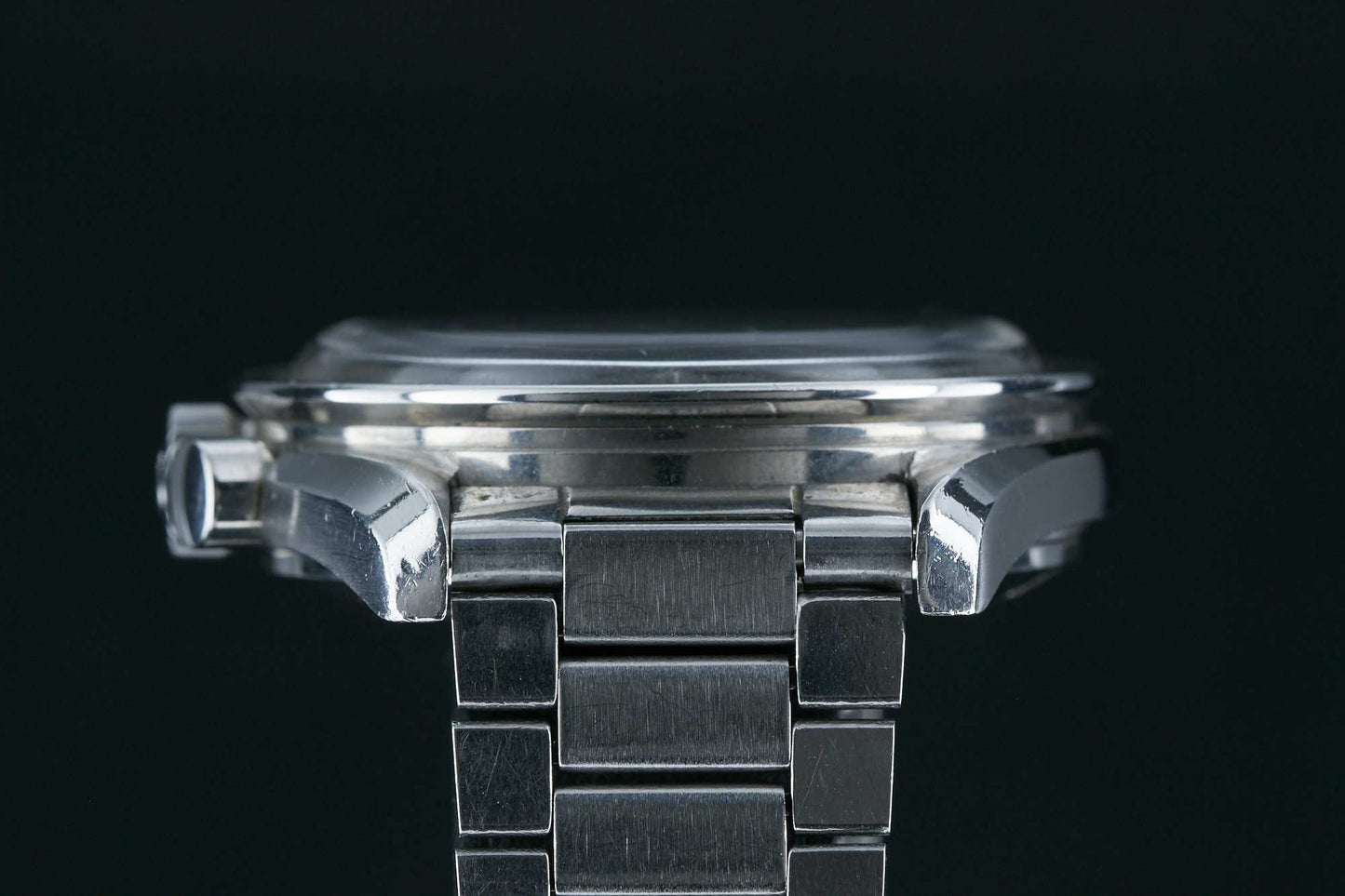 Close-up side view of the Omega Speedmaster Professional by Omega, a silver metal wristwatch with a link bracelet, highlighting the case, bezel, and part of the crown against a black background to showcase its profile and construction details.