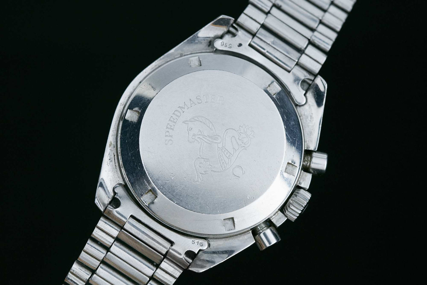 The back of an Omega Speedmaster Professional chronograph with a metal bracelet features engraved "Speedmaster" text and a seahorse image on the case back.