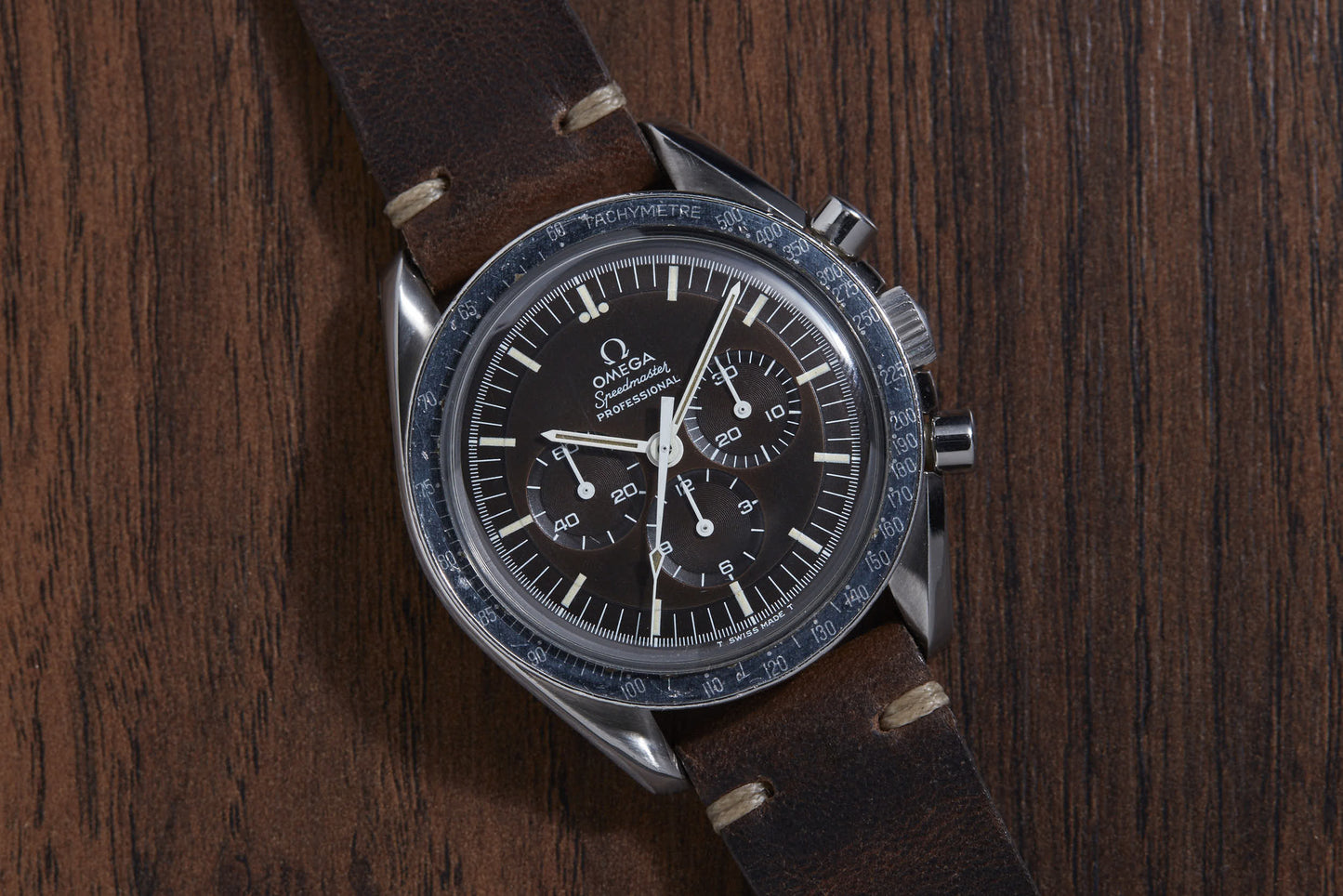 An Omega Speedmaster Professional wristwatch by Omega, featuring a black dial with three subdials and a tachymeter bezel, is shown on a brown leather strap resting on a wooden surface. Famously associated with the Moon Landing.