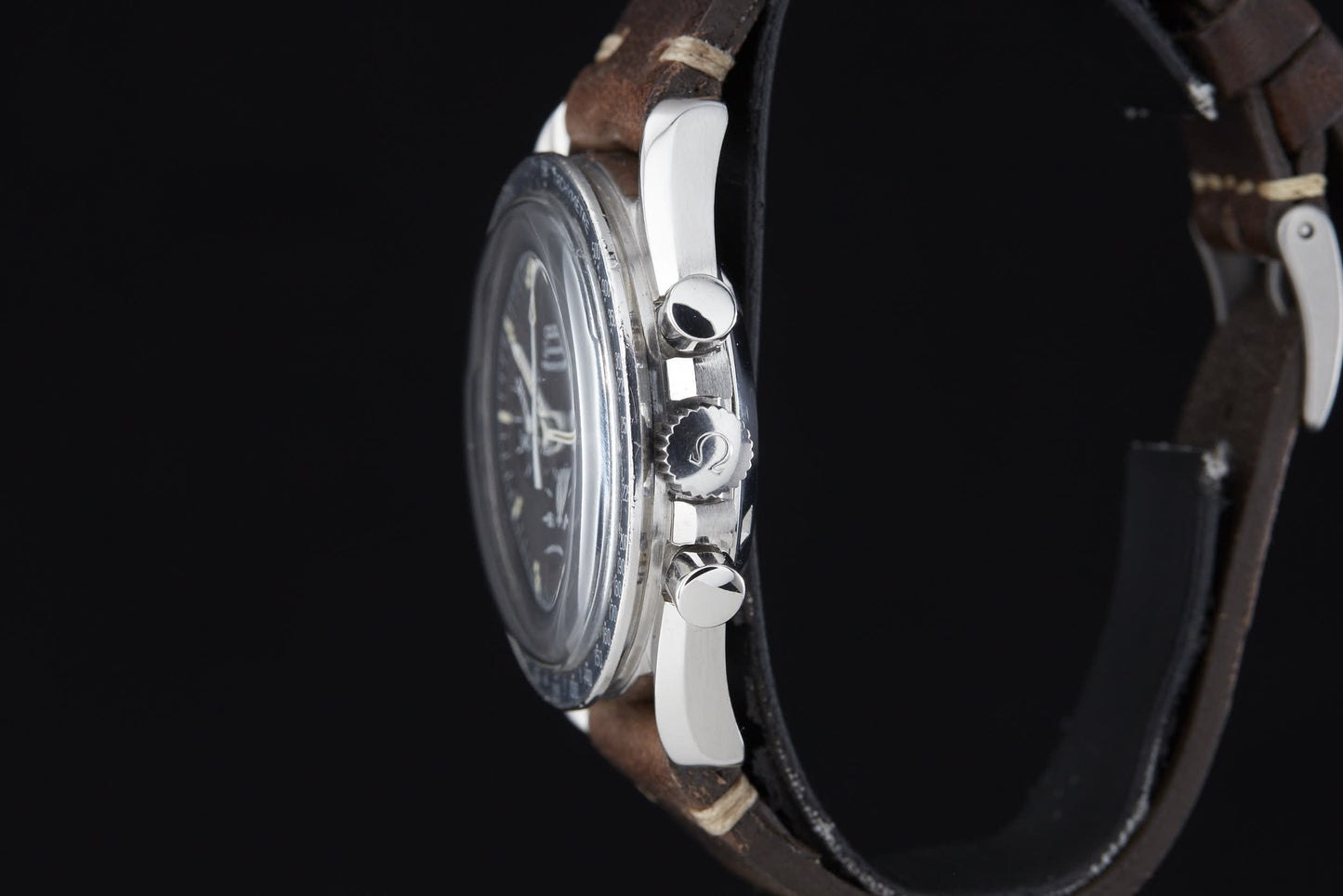 A close-up side view of the Omega Speedmaster Professional by Omega, featuring a metal case and brown leather strap reminiscent of the Apollo era, with crown and pushers highlighted against a black background.