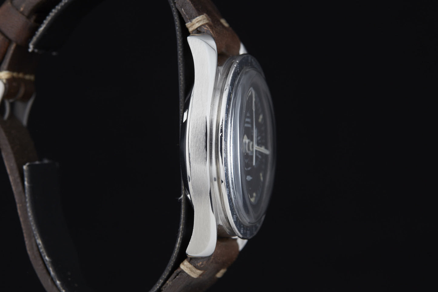 Close-up side view of the Omega Speedmaster Professional by Omega—brushed silver case, clear glass, and brown leather strap with visible stitching—timeless elegance inspired by Apollo ingenuity, set against a black background.