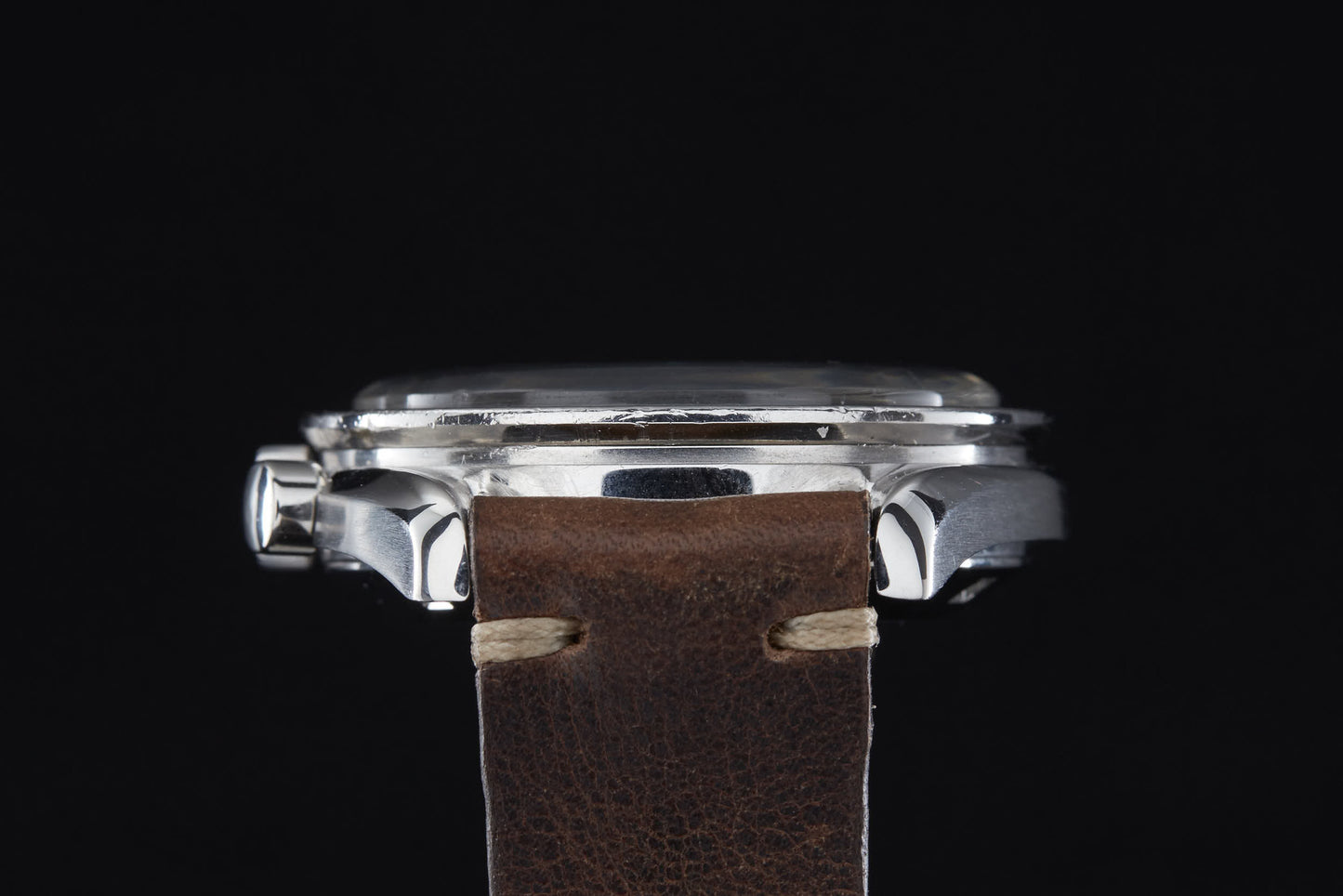 A close-up side view of the Omega Speedmaster Professional by Omega, featuring a silver case and brown leather strap against a black background, with crown and stitching details—an elegant tribute to Apollo-era innovation.
