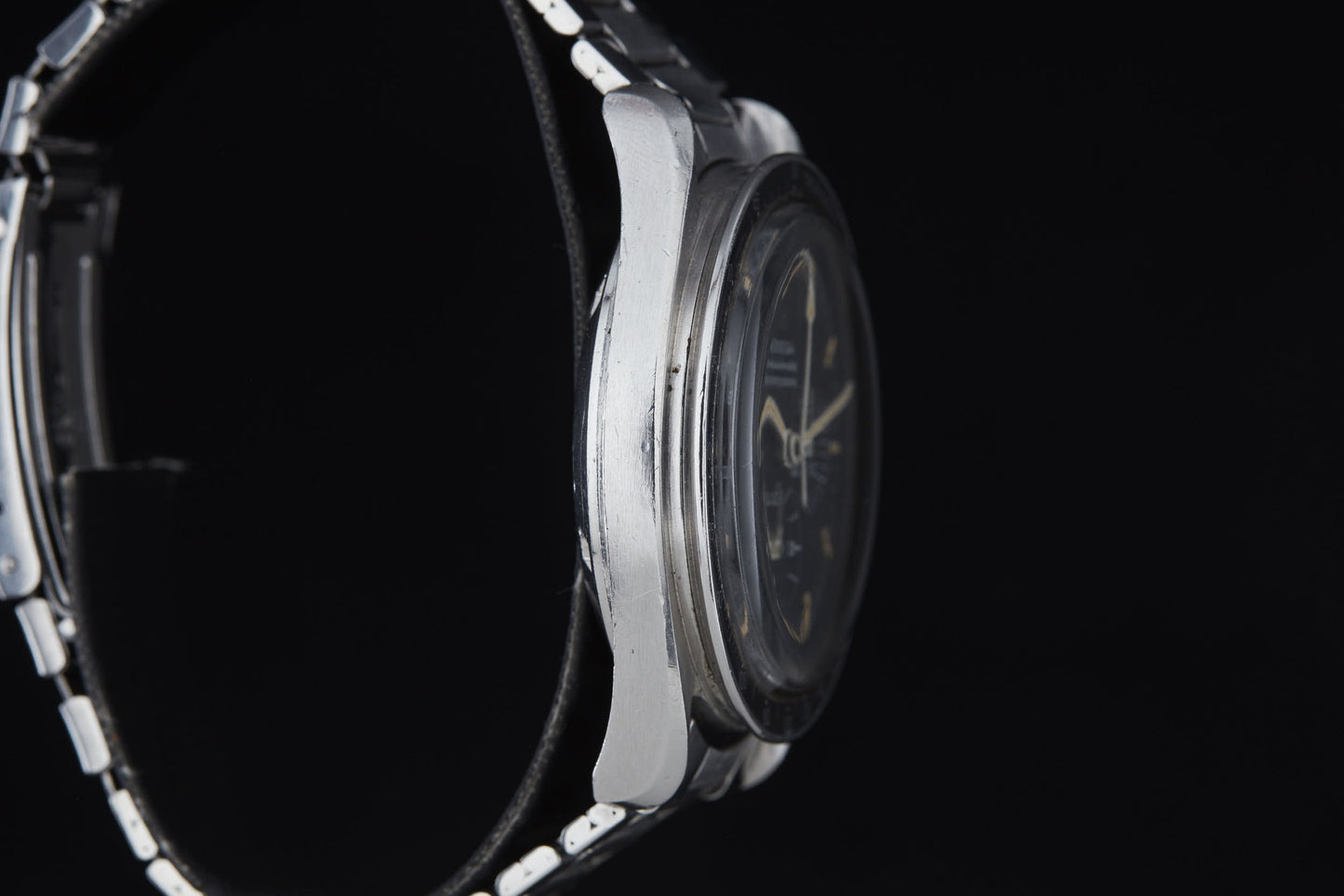 Close-up side view of a silver Omega Speedmaster Professional by Omega with a metal band, displaying wear and scratches on the case against a black background. The partially visible watch face is seen through its domed crystal, echoing its Apollo mission legacy.