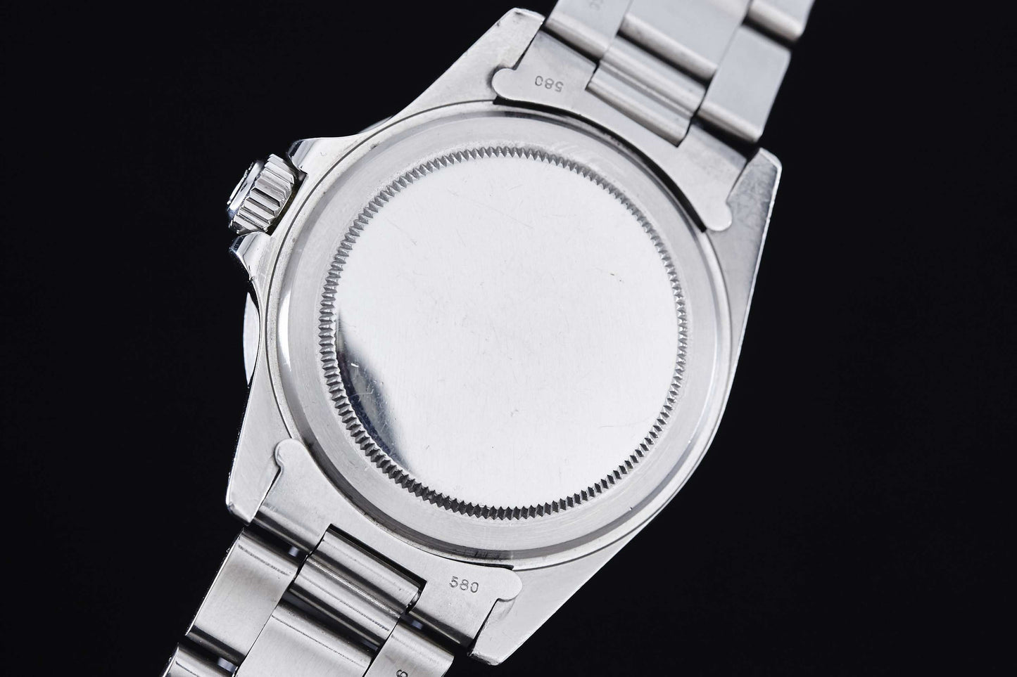 The image shows the back of a Rolex "Red" Submariner stainless steel dive watch with a metal bracelet. Its smooth, round case back has a knurled edge for opening, no visible markings, and the crown is on the left side.