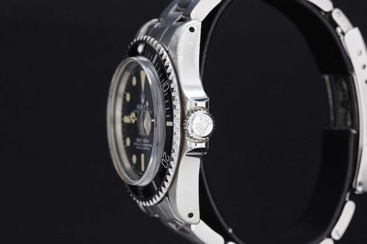 Close-up side view of the Rolex "Red" Submariner dive watch, featuring a black bezel and dial, silver case with logo-engraved crown, and a partially visible black strap against a black background.
